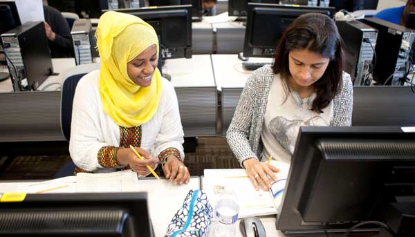 Two female students in front of computers looking at a book
